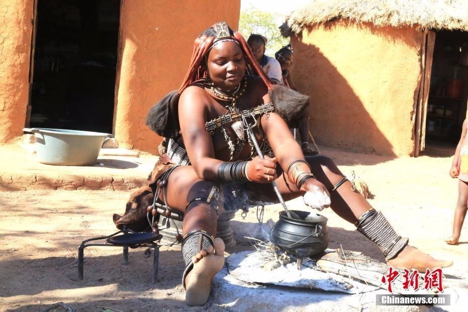 生活在纳米比亚的辛巴族又称“红泥族”，是非洲最后保持原始生态的部落之一。作为该部落最大特色，辛巴族女子终生不洗澡，且常年袒露上身，尤其喜欢把一种红色矿物粉末混合着奶油等涂抹在身上和头上。图为“红泥族”人在房屋前生火做饭。中新社记者 王曦 摄